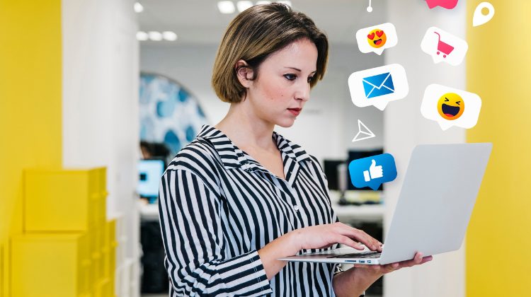 Mulher usando notebook com ícones de redes sociais ao redor, simbolizando trabalho com mídias digitais.