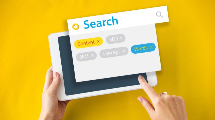 Mãos segurando um tablet com tela mostrando uma interface de pesquisa com palavras-chave relacionadas a SEO e marketing digital.